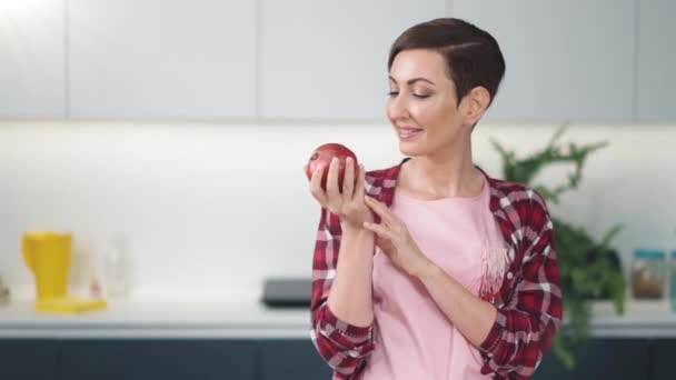 Hemmafru håller färskt äpple i handen bär rutig skjorta med en kort frisyr medan matlagning äppelpaj står vid köket. Friska livsmedel hemma. FHD-bilder — Stockvideo
