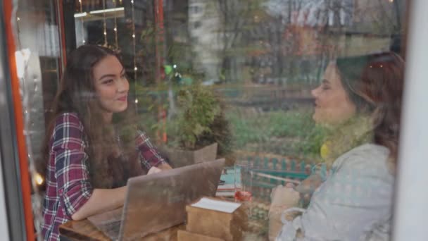 Widok przez szybę dwie uczennice rozmawiają przy kawie razem w kawiarni z klientami chodzącymi po tle, gdy siedzą przy stole i rozmawiają na laptopie. Materiał 4K — Wideo stockowe