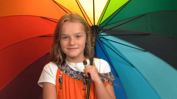 Charmantes kleines Mädchen in orangefarbenem Kleid hält Regenbogenfarben Regenschirm stehend und jubelnd im Studio, während sie in die Kamera schaut — Stockfoto