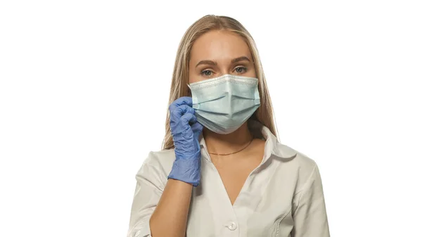 Colocando em uma máscara médica rosto jovem enfermeira loira olhando para a câmera vestindo uniforme médico branco e brilho de borracha azul descartável isolado no fundo branco — Fotografia de Stock