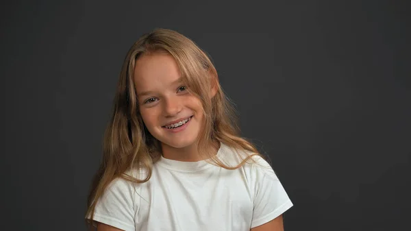Glimlachend meisje met strik zijwaarts hoofd kijkend naar de camera in wit t-shirt geïsoleerd op donkergrijze of zwarte achtergrond. Het begrip "kinderemoties" — Stockfoto