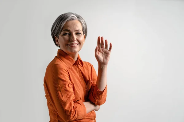 Grauhaarige hübsche Frau mittleren Alters lächelt sanft in die Kamera, mit anrührenden Fingerspitzen in orangefarbenem Hemd und Jeans auf weißem Hintergrund. Konzept menschlicher Emotionen — Stockfoto