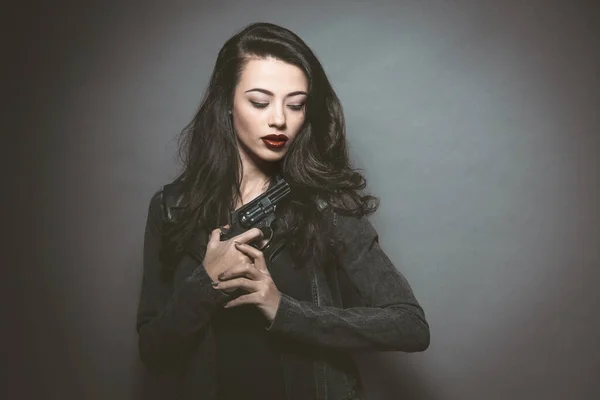 Agente secreto bela jovem com lábios vermelhos segurando uma arma usando jaqueta de couro. Modelo feminino jovem perigoso com maquiagem profissional. Moda, beleza, maquiagem, penteado e arma — Fotografia de Stock