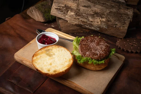 Fresh craft beef burger with cherry sauce in a sauce bowl, fresh lettuce with lead bun laying next with wooden lodges as decoration. Restaurant concept. Street food concept — Stock Photo, Image