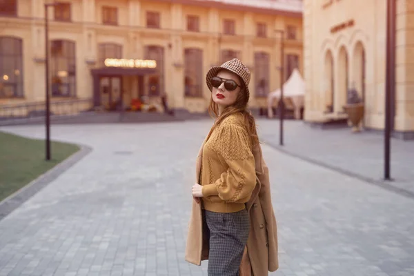 Tourné de derrière mannequin jeune femme en lunettes de soleil et accroché sur une épaule automne manteau beige marchant dans la rue posant joyeusement pour la caméra. Photo teintée — Photo