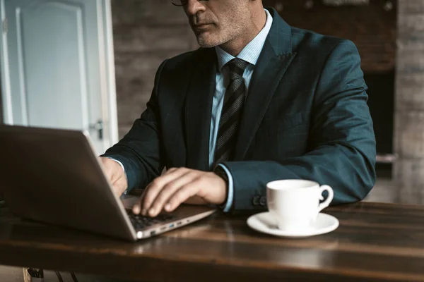 Brak widocznego biznesmena siedzącego przy stole, pracującego nad laptopem i pijącego kawę w nowoczesnym biurze. Człowiek pracujący na laptopie w biurze. Koncepcja biznesowa i finansowa — Zdjęcie stockowe