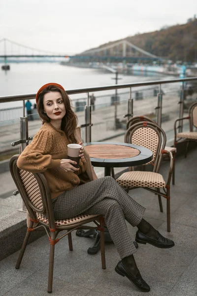 Sentado de lado convertido mujer joven parisina en la terraza del restaurante. Retrato de una mujer joven y elegante con abrigo de otoño y boina roja al aire libre —  Fotos de Stock