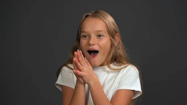 Spännande liten flicka står med händerna ihop glatt ser en sida av kameran bär vit t-shirt isolerad på mörkgrå eller svart bakgrund — Stockfoto