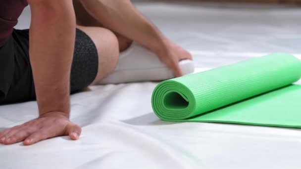 Hombre atlético relajante sentado en el suelo junto a la alfombra de rollo desenrollado, lugar de limpieza después de los deportes. Hombre deportivo haciendo entrenamiento físico. Un joven fuerte en el gimnasio. Imágenes 4K — Vídeo de stock