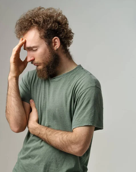 Hat Kopfschmerzen oder Scham für einen gegnerischen jungen Mann, der seine Stirn berührt. Schöner junger Mann mit lockigem Haar in olivem T-Shirt auf weißem Hintergrund — Stockfoto