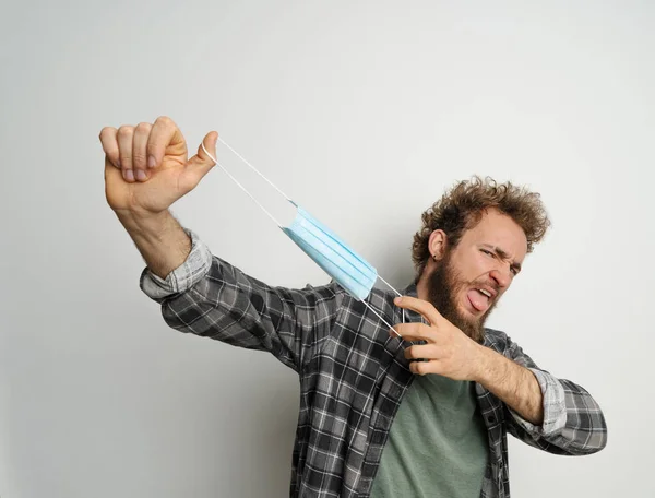 Played or got rid of young man holding protective medical mask in front of face ready to end quarantine, with curly hair, wearing plaid shirt and olive t-shirt under. White background — Stock Photo, Image