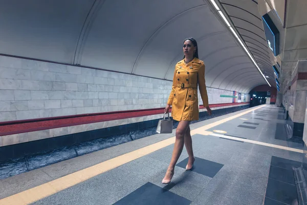 Marcher sur la plate-forme de métro belle jeune fille avec de longues belles jambes dans un manteau de printemps jaune et un sac à main blanc ou sac à main à la main — Photo