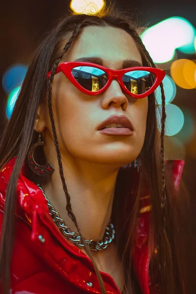 Outdoor modeportret van glamour jonge vrouw met gevlochten haar draagt rood naar beneden jas en mode rode zonnebril op neon straatverlichting. Nachtclub mode — Stockfoto