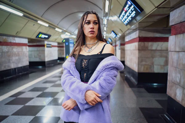 Chica modelo de moda en abrigo de piel sintética púrpura de pie desnudó sus hombros en la plataforma de paso del metro caminando. Mujer con maquillaje de lujo, pelo largo en traje elegante — Foto de Stock