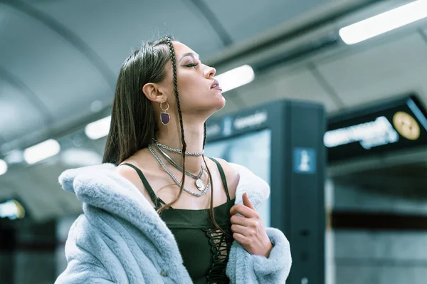 Model busana wanita muda dengan mantel bulu sintetis ungu berjalan di metro atau platform kereta bawah tanah. Perempuan dengan riasan mewah, rambut panjang dalam gaya pakaian — Stok Foto