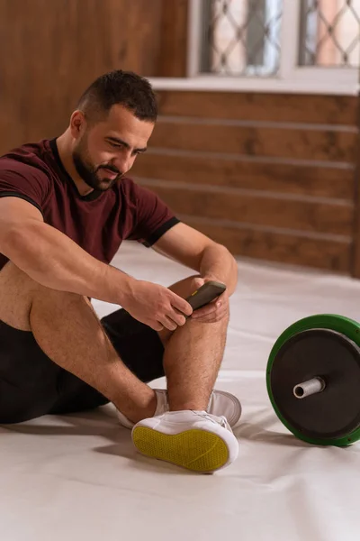 Hombre guapo sentado en un piso con teléfono inteligente en sus manos y barra de fitness de tono negro y verde, equipo para el concepto de entrenamiento con pesas. Equipamiento deportivo para entrenamiento. concepto de estilo de vida saludable — Foto de Stock