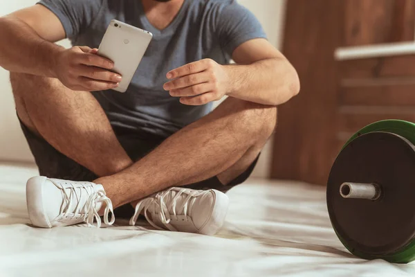 Ningún joven visible de la cara que tiene una rotura que se sienta en un piso con la barra negra y verde de la aptitud del tono, equipo para el concepto del entrenamiento con pesas. Equipamiento deportivo masculino para entrenamiento — Foto de Stock