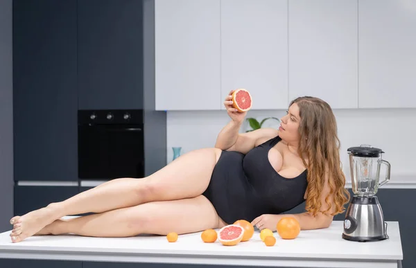 Sexy fille en surpoids allongé sur la table de cuisine. Une jeune fille blanche potelée sexy obèse en maillot de bain noir dans la cuisine moderne avec des fruits à côté d'elle — Photo