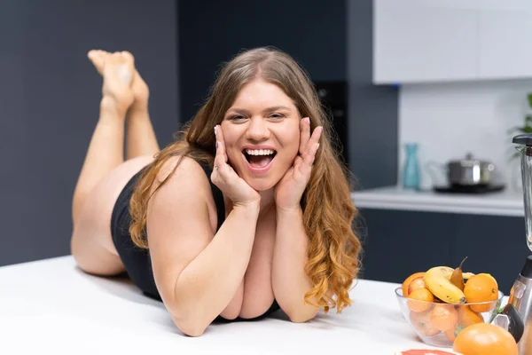 Overweight girl with big breasts laying on the kitchen table. An obese young sexy chubby white girl in black swimsuit at modern kitchen with fruits next to her — Stock Photo, Image