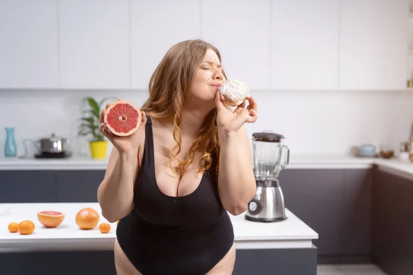 Επιλέγοντας υγιή ή ανθυγιεινά τρόφιμα θετική γυναίκα στέκεται στην κουζίνα κρατώντας γκρέιπφρουτ και daughnut με πολλά φρούτα στο τραπέζι δίπλα στο blander. Σέξι υπέρβαρο κορίτσι κάθεται στο τραπέζι της κουζίνας — Φωτογραφία Αρχείου