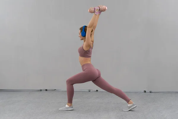 Stretching using dumbbells lifted hands up young girl losing weight at home wearing pink outfit while listen music in blue wireless headphones. Sport and recreation concept — 图库照片