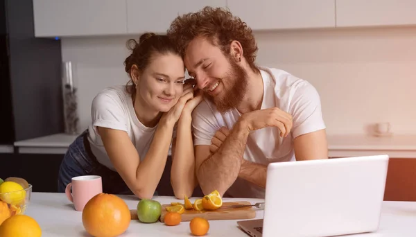 Vackra unga par pratar på videosamtal med bärbar dator. Unga par matlagning hälsosam mat i köket hemma. Mannen lutade sig mot flickan leende tittar på romantisk film — Stockfoto