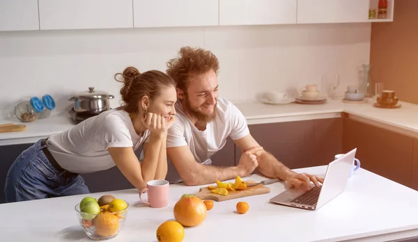 Νεαρό ζευγάρι μαγειρεύει υγιεινό φαγητό στην κουζίνα στο σπίτι. Το κορίτσι έσκυψε πάνω στον άντρα που χαμογελούσε βλέποντας ρομαντική ταινία. Όμορφο νεαρό ζευγάρι μιλάει σε βιντεοκλήση χρησιμοποιώντας το laptop — Φωτογραφία Αρχείου
