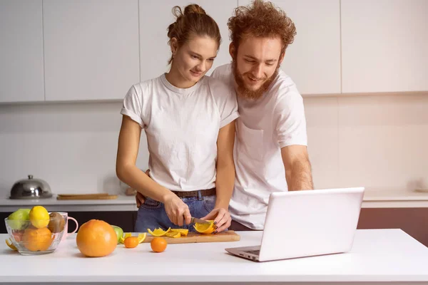 Homme appuyé sur fille souriant regarder un film romantique. Beau jeune couple parlant sur appel vidéo à l'aide d'un ordinateur portable. Jeune couple cuisine des aliments sains dans la cuisine à la maison — Photo