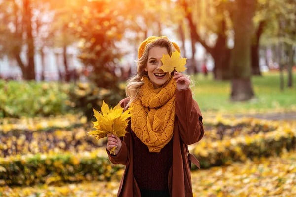 Щаслива жінка з жовтим листом у жовтому в'язаному береті з осіннім листям в руках і осіннім жовтим садом або парком. Красива усміхнена молода жінка в осінньому листі — стокове фото