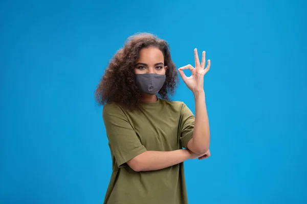 Dizendo OK usando as mãos para se comunicar muito afro-americano menina em máscara protetora reutilizável. Curly cabelo jovem mulher mostrando Olá sinal falando na câmera isolada no fundo azul — Fotografia de Stock