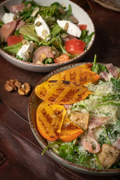 Dos tazones de ensaladas con salmón, queso mozzarella y rebanadas de frutas a la parrilla están sobre una mesa cubierta con un trapo de cuero. Concepto de restaurante. Imagen vertical —  Fotos de Stock