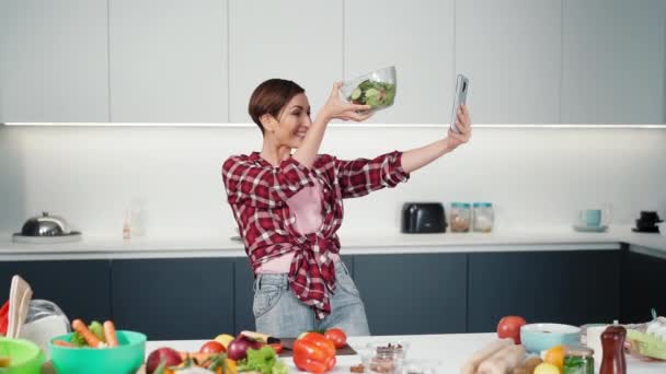 Felice casalinga fare selfie con il suo smartphone in possesso di un bawl di insalata fresca fatta in mano tesa, mentre si cucina in cucina moderna a casa indossando una camicia a quadri. Al rallentatore. Filmati FHD — Video Stock