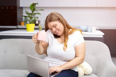 Drinking coffee or tea while working on laptop sitting at home curve body young woman staying at home during quarantine. Self isolation as prevention. Working distantly from home using laptop clipart