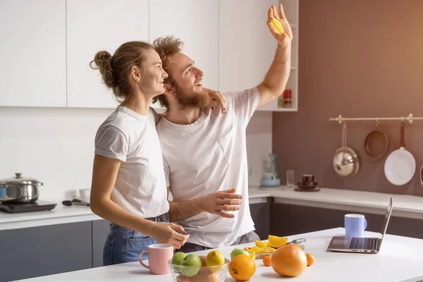 Κρύβονται από μια ακτίνα φωτός που λάμπει από ένα παράθυρο νεαρό ζευγάρι κάνει πρωινό στην κουζίνα στο σπίτι. Κορίτσι αγκαλιά σύζυγος χαμογελά κοιτάζοντας μακριά. Όμορφο νεαρό ζευγάρι στην κουζίνα — Φωτογραφία Αρχείου