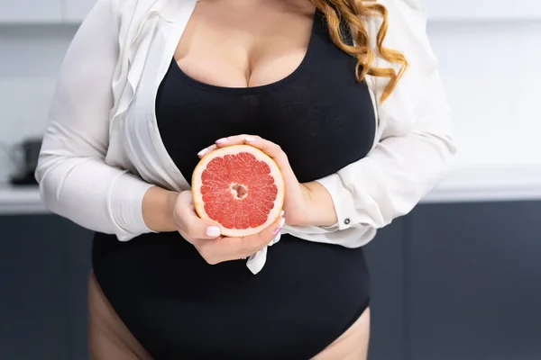 Закройте изогнутое тело молодой женщины, держащей в руках половину свежего грейпфрута, стоящего дома на современной кухне с длинными светлыми волосами. Диета и питание. — стоковое фото