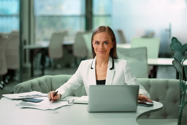 Mulher de negócios bonita loira trabalhando com documentos sentados na frente do laptop vestindo terno oficial branco. Trabalhador de escritório olhando para a câmera com sorriso enquanto está sentado em seu local de trabalho — Fotografia de Stock