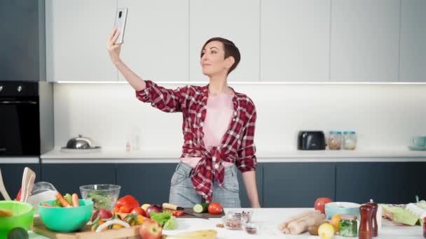 Jeune blogueuse culinaire joyeuse utilisant son smartphone à la main tendue, tout en cuisinant dans la cuisine moderne à la maison portant une chemise à carreaux. Au ralenti. Vidéos FHD — Video