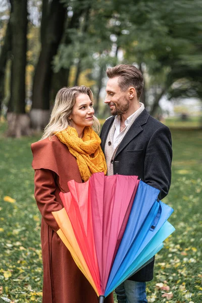 Ser in i ögonen på varandra med stängd regnbåge paraply vacker i kärlek par som står i parken under en. Ett vackert par i höstparken i regnigt väder — Stockfoto