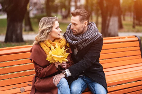 Glada kärleksfulla par omfamna sitter på bänken romantisk kramas i parken bär rockar och halsdukar Samla en bukett fallna blad. Kärlekshistoria. Tonad bild — Stockfoto