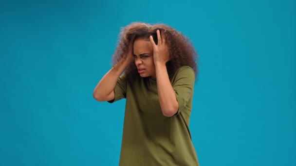 Zestresowana lub nieszczęśliwa Afroamerykanka dotykająca głowy cierpiąca na ból głowy, nosząca koszulkę z oliwek odizolowaną na niebieskim tle. Koncepcja piękna. Koncepcja opieki zdrowotnej — Wideo stockowe