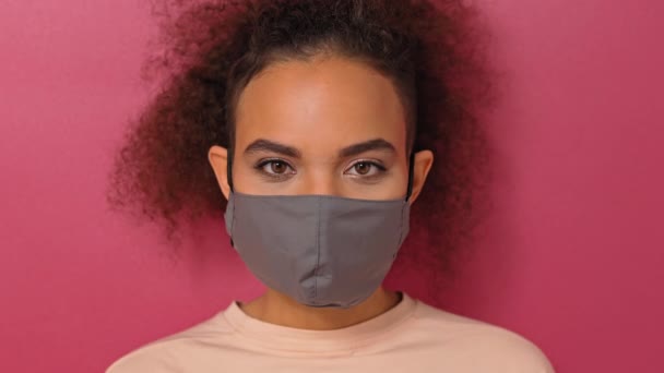 Przesłuchanie lub studiowanie wygląd noszący maskę wielokrotnego użytku Afroamerykanin uroczy dziewczyna w ślicznej t-shirt, aby zapobiec innym od korony COVID-19 i zakażenia SARS cov 2 izolowane na różowym tle — Wideo stockowe