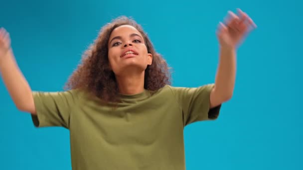 Komik pilot hareketleriyle komik genç kız Afro-Amerikalı kadın kameraya olumlu bakıyor şeftali rengi tişört giyiyor ve mavi arka planda izole edilmiş kaslarını gösteriyor. 4K görüntü — Stok video