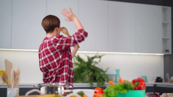 Die junge Hausfrau mit den kurzen Haaren tanzt in der Küche und kocht mit einem langen Kochlöffel, der in der modernen Küche steht, ein Abendessen für die Familie. FHD-Filmmaterial — Stockvideo