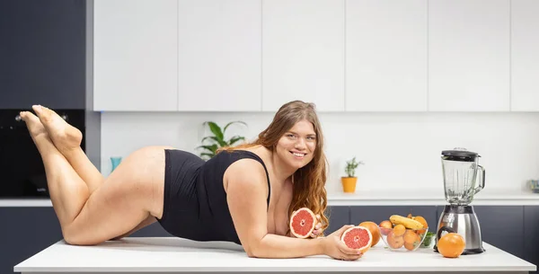 Sexy corpo positivo paffuto ragazza sdraiata sul tavolo della cucina indossando costume da bagno nero in possesso di un pompelmo fresco in mano. Ragazza grassa con lunghi capelli biondi ricci. Dieta e nutrizione concetto — Foto Stock