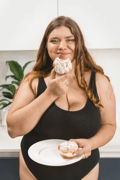 Satisfait jeune femme grosse mangeant un gâteau heureux souriant à la caméra portant un maillot de bain noir. Belle jeune femme joufflue qui mange des aliments malsains. Fat girl manger gâteau debout sur la cuisine moderne — Photo
