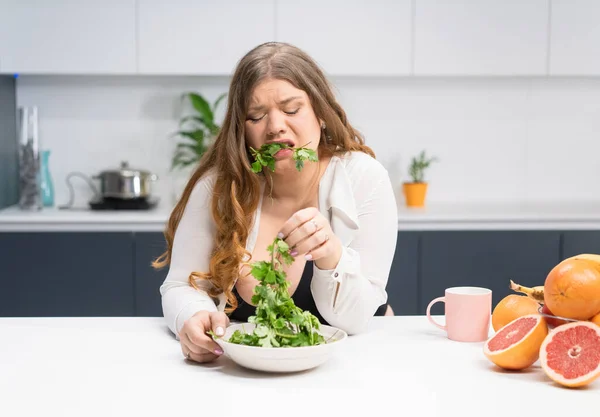 Gadis muda dengan masalah berat badan memegang segar salad casting mencoba untuk mengunyahnya. Wanita muda bertubuh melengkung dengan rambut pirang panjang duduk di dapur modern. Konsep diet dan nutrisi — Stok Foto