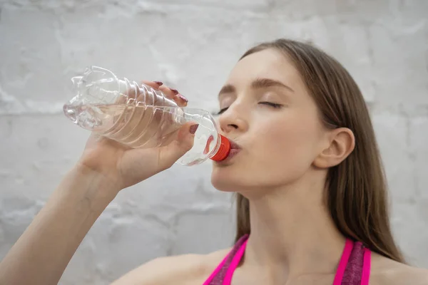 Joven atleta mujer beber agua de botella de plástico mientras se toma un descanso. Hermosa chica atlética En color rosa entrenamiento deportivo ropa de fitness en la pared blanca Fondo. Imagen de alta calidad —  Fotos de Stock