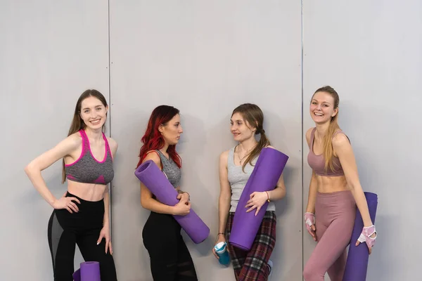 Meninas atléticas em esportes se encaixa em pé ao lado da parede segurando um tapete de ioga, sorrindo na câmera. Praticando no estúdio de fitness ou ioga, exercitando-se no interior. Isolado sobre fundo cinzento — Fotografia de Stock