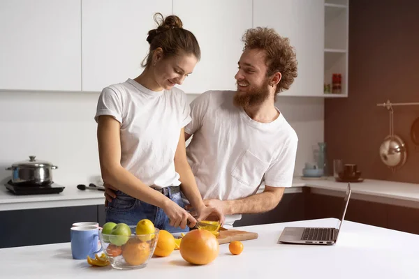 Ευτυχισμένος από το νέο τους σπίτι νεαρό ζευγάρι μαγείρεμα υγιεινό φαγητό στη νέα κουζίνα βλέποντας online streaming βίντεο στο laptop. Όμορφο νεαρό ζευγάρι μιλάει σε βιντεοκλήση χρησιμοποιώντας το laptop. Νέα αρχική ιδέα — Φωτογραφία Αρχείου