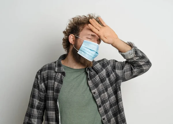 Sentindo o jovem de alta temperatura usando máscara médica estéril protetora em seu rosto para proteger o cabelo encaracolado do coronavírus na camisa xadrez e na camiseta de oliva embaixo. Conceito de medicina. Fundo branco — Fotografia de Stock